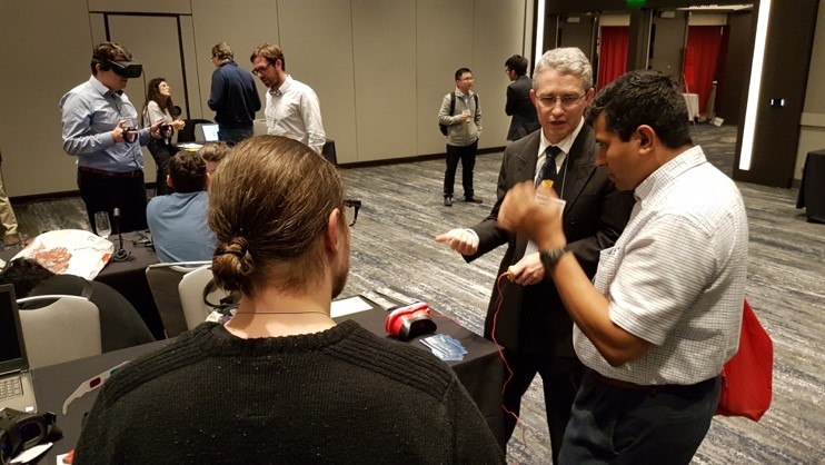 Figure 3. The demo session is always accompanied by a number of interesting discussions. Moreover, this photo contains three Google VR cardboards / HMDs  find them all!  In the mid-frame is Andrew Woods in discussion with Guarav Sharma.